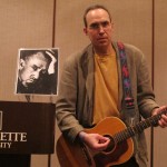 Paul strums his acoustic guitar on stage beside a photo of Steinbeck.